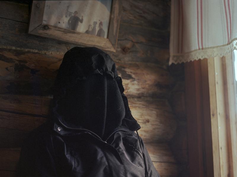 An unknown figure, in a black winter jacket with a hood and a thick black fabric covering the face inside the hood. Sitting in a smoky old log cabin.